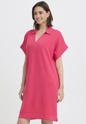 Mosolygó nő laza, térdig érő, élénk rózsaszín ruhában, amely galléros és rövid, feltűrt ujjú, egy egyszínű háttér előtt áll.