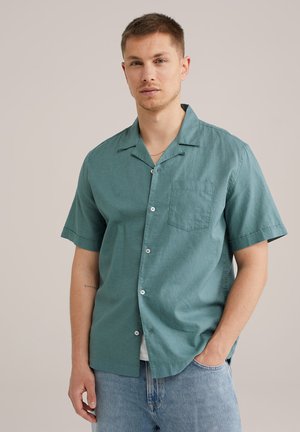 Chemise à manches courtes en tissu teal avec une poche avant. Présente un design à boutons et une texture douce, associée à un jean bleu clair.