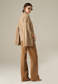 Maglione beige a maglia con maniche ampie, abbinato a pantaloni brown a gamba larga. Il modello indossa occhiali da sole oversize e orecchini a goccia.