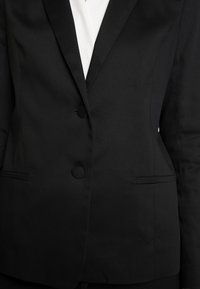 Blazer negro ajustado con una textura suave, solapas con muesca, dos botones y bolsillos laterales, llevado sobre una blusa blanca.