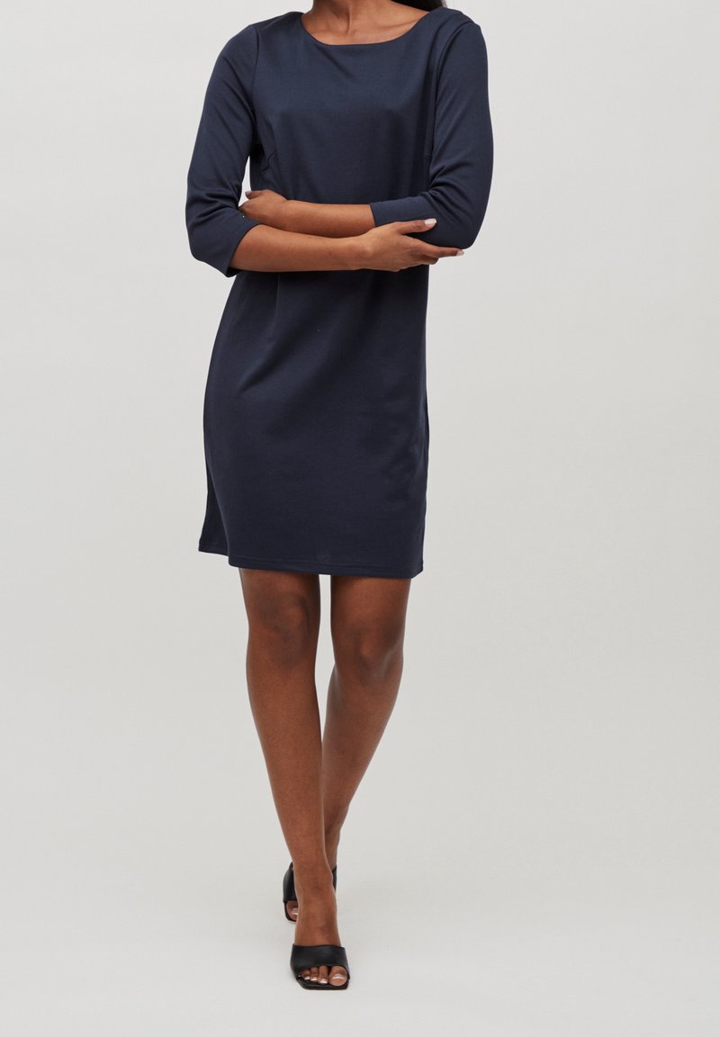 Femme en robe bleu marine longueur genoux avec manches trois-quarts, debout les bras croisés et portant des talons ouverts noirs.
