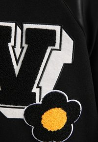 Prenda negra con una letra "V" blanca y esponjosa, y una flor negra texturizada con un centro amarillo. Diseños audaces con costuras en contraste.