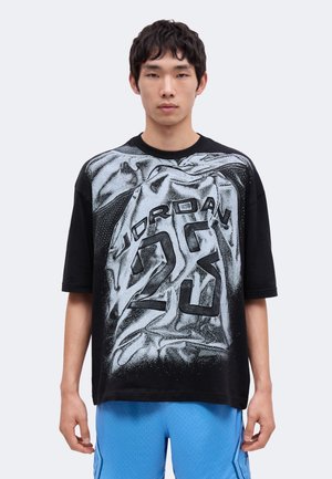Junger Mann trägt ein schwarzes oversized T-Shirt mit einem großen weißen Aufdruck eines Jordan-Basketballtrikots mit der Nummer 23, kombiniert mit blauen Shorts.