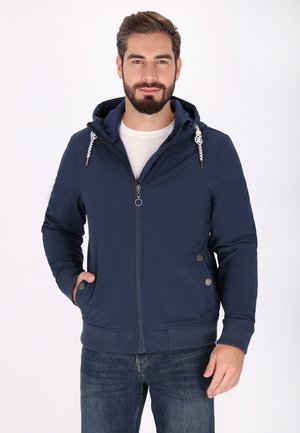 Man with beard wearing a navy blue zip-up hoodie with drawstrings and blue jeans, standing with one hand in pocket against white background.