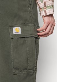 Gröna cargobyxor tillverkade av slitstarkt tyg, med en stor sidficka och en fyrkantig logopatch i gult och rött på den främre fickan.