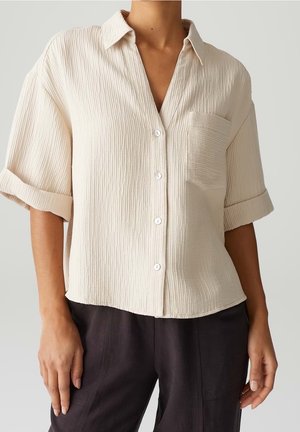 Femme portant une chemise crème texturée à boutons avec manches courtes retroussées et pantalon noir sur un fond uni.