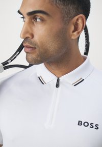 White polo shirt with a zippered collar, featuring a textured fabric, black and brown striped accents, and a "BOSS" logo on the front.