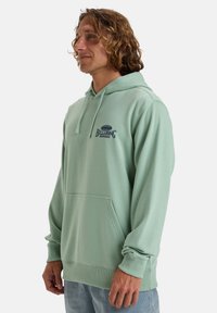 Sudadera con capucha de tela verde claro con un bolsillo canguro en la parte frontal. Presenta un logo en azul oscuro en el pecho. Textura suave, corte casual.