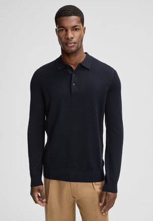 Man draagt een marineblauw, gestructureerd, lang mouw poloshirt en beige broek, staand tegen een effen lichtgrijze achtergrond.