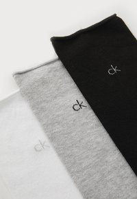 Kolm kokku volditud Calvin Kleini sokki valges, hallis ja mustas, igaühel on väike "ck" logo lähistel.