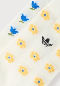 Hvite sokker med broderte blå blomster med grønne stilker og gule blomster, samt en liten svart fuglelogo. Strukturert strikket stoff.