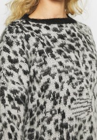 Weicher, flauschiger Strickpullover mit schwarz-grauem Leopardenmuster und subtilen Glitzerelementen an einer Person mit blonden Haaren.