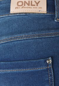 Blå denimjeans med en slät textur, utrustade med gyllene sömmar och en märkesläderlapp vid midjan. Liten metallknapppunkt synlig.