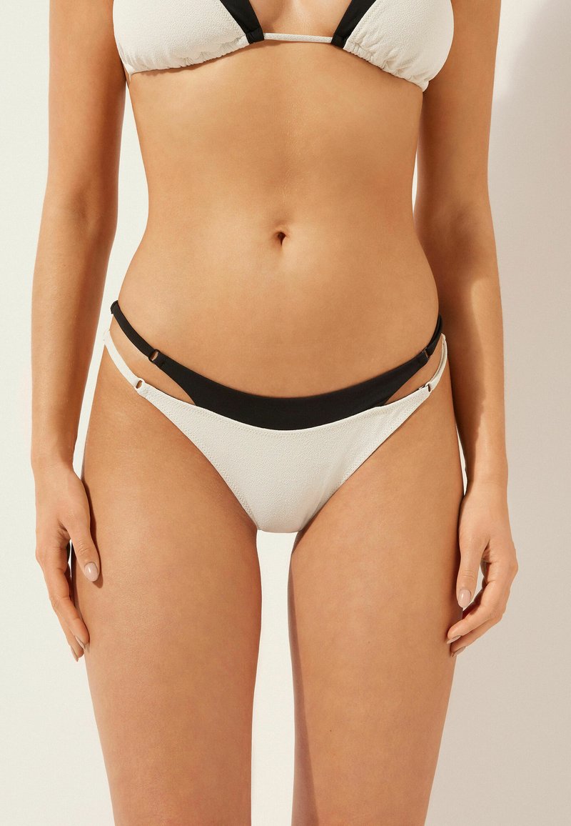 Komplet bikini z teksturowanym beżowym topem i czarnymi ramiączkami, z regulowanymi bocznymi wiązaniami oraz niskim krojem dolnej części. Zawiera minimalistyczne akcenty.