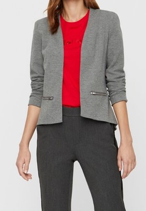 Vrouw die een grijs getailleerd colbert met ritspakken draagt over een rood shirt en donkergrijze, op maat gemaakte broek, staand tegen een effen achtergrond.