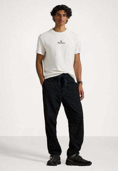 Λευκό βαμβακερό μπλουζάκι με μαύρο λογότυπο "Ralph Lauren", σε συνδυασμό με μαύρες παντελόνες με κορδόνι και μαύρα αθλητικά παπούτσια. Το μοντέλο στέκεται με τα χέρια στις τσέπες.