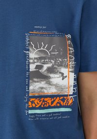 Niebieska bawełniana koszulka z grafiką przedstawiającą słońce, scenę skateboardową oraz stylizowany tekst w kolorze pomarańczowym i białym. Krótkie rękawy.