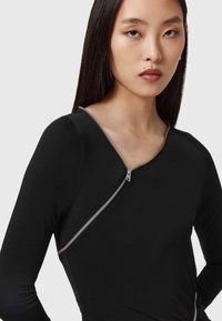 Svart langermet topp med en skrå sølvglidelås, tettsittende design og glatt tekstur med rynkede detaljer på siden.