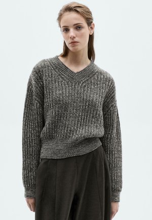 Young woman with light hair wearing a gray textured V-neck sweater and dark wide-leg trousers, standing against a plain light background.