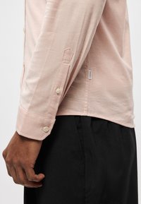 Ροζ πουκάμισο με μακριά μανίκια και απαλή υφή, με μανσέτες με κουμπιά και ετικέτα στο πλάι της ραφής. Φοριέται πάνω από μαύρο παντελόνι, αναδεικνύοντας τις λεπτομέρειες των μανικιών.