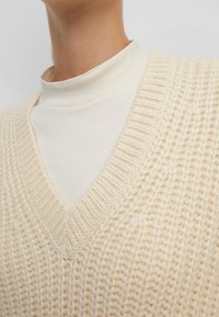 Pull col en V en maille crème superposé à un col roulé crème. Détail côtelé visible au niveau de l'encolure et de l'ourlet du pull.