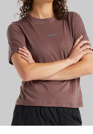 Kobieta w brązowej koszulce z krótkim rękawem z logo "icebreaker", z założonymi rękami, ubrana w czarne spodnie, na jednolitym szarym tle.