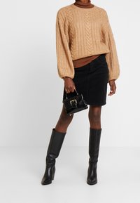 Beige gebreide trui met ribbelstructuur, zwarte corduroy rok, zwarte kniehooglaarzen en een kleine zwarte handtas met een sluiting.
