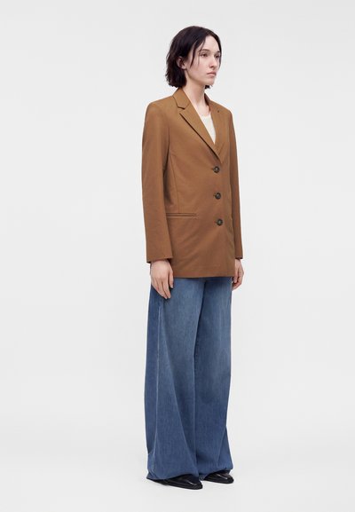 Femme debout portant un blazer marron boutonné sur un haut clair avec un jean bleu à jambes larges et des chaussures noires pointues sur un fond clair uni.