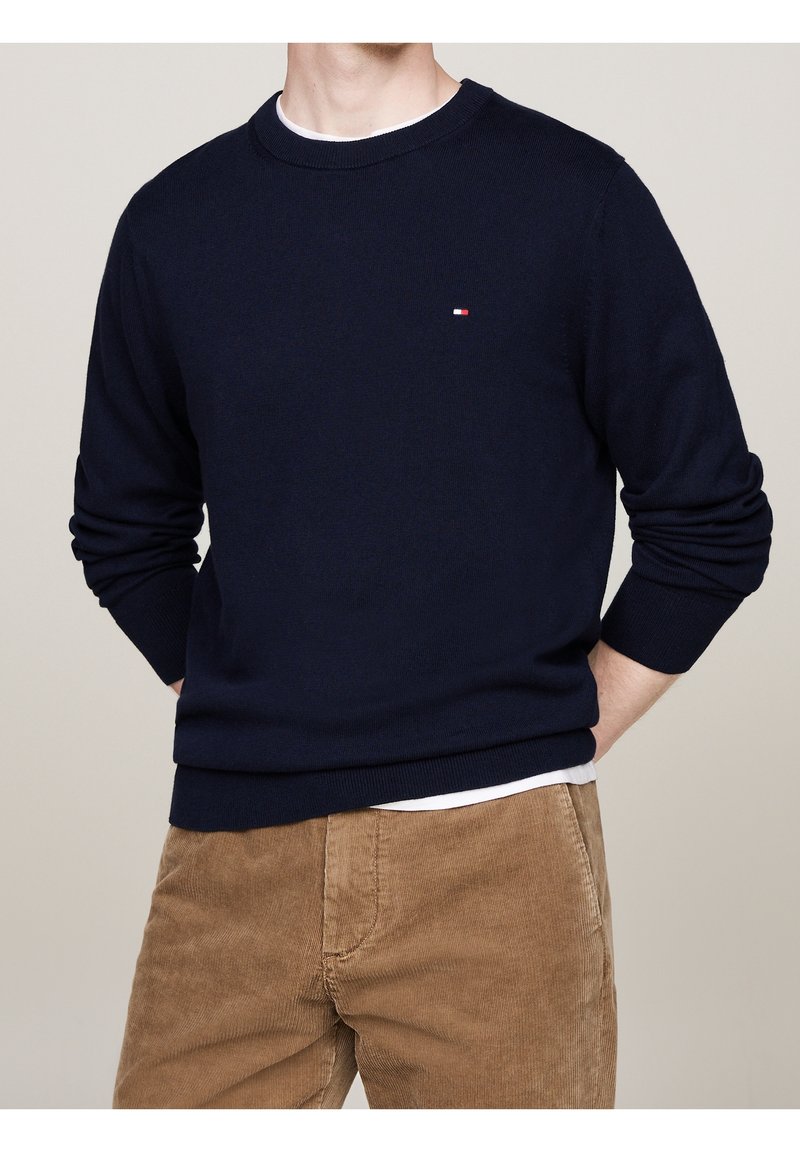 Dunkelblauer Strickpullover mit Rundhalsausschnitt und langen Ärmeln, mit einem kleinen rot-weißen Logo auf der linken Brust. Kombiniert mit braunen Cordhosen.