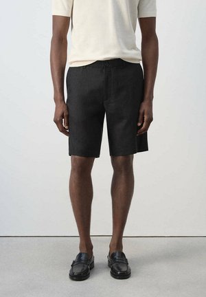 Modelo masculino con pantalones cortos negros a medida, camisa blanca de manga corta y mocasines de cuero negros, de pie frente a un fondo claro y liso.