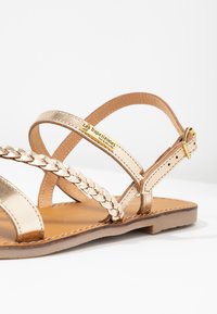 Sandali metallici dorati con cinturini intrecciati a incrocio, suola piatta e una piccola chiusura con fibbia. Etichetta del marchio visibile sul cinturino laterale.