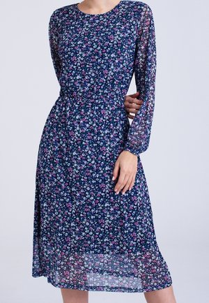 Kvinna iklädd en knälång marinblå klänning med långa skira ärmar och ett rosa, vitt och blått blommönster mot en enfärgad bakgrund.