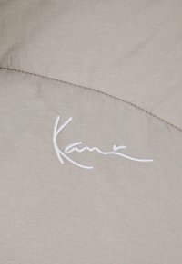 Tela gris claro con una firma en bordado blanco, con detalles de costura visibles que crean una apariencia texturizada.