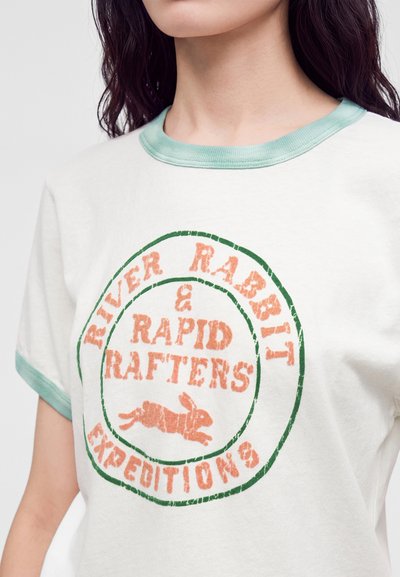 T-shirt blanc avec bordure vert clair porté par une personne, avec un logo circulaire représentant un lapin et un texte indiquant "River Rabbit & Rapid Rafters Expeditions."