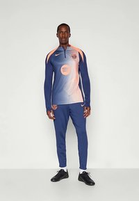 Vīrietis modelis, tērpies zilā un oranžā FC Barcelona treniņtērpā un biksēs ar melnām sporta kurpēm, stāvot pret vienkāršu baltu fonu.