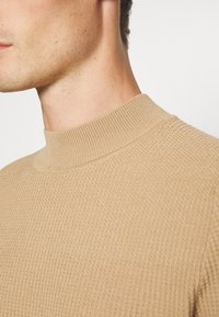 Beige gestrickter Pullover mit einem hohen, gerippten Kragen. Die Textur weist ein dezentes Waffelmuster auf, das das leichte Material betont.
