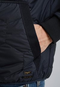 Marinefarbener, gesteppter Mantel mit sechseckigem Muster, schwarzem Besatz und einer seitlichen Tasche mit einem Etikett, auf dem "Americana Classic, Typ Nr.: C-46" steht.