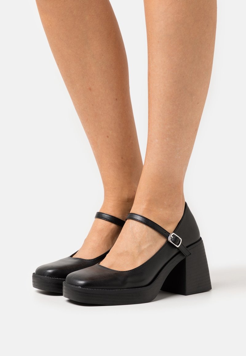 Talons compensés noirs avec un bout rond, une bride de cheville et un talon carré. Texture en cuir lisse avec une légère brillance et un détail de boucle en argent.