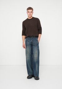 Ung man står med en hand i fickan iförd en mörkbrun tröja, vida blå jeans med lappar, svart bälte och svarta sneakers.