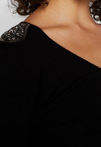 Pull noir ajusté avec un large décolleté. L'épaule présente des perles décoratives et un contraste de textures par rapport au tissu lisse.