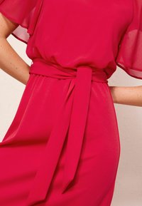 Gros plan d'une personne portant une robe rouge vif avec des manches flottantes transparentes et une ceinture en tissu nouée à la taille.