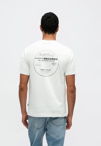 Valge puuvillane t-särk, mille seljal on vinüülplaadi graafika tekstiga: �„Boss Club Records, The Hamburg Sound.“ Lahtine lõige.