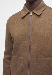 Gros plan d'une personne portant une veste marron zippée avec un col et deux poches avant.