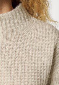 Gros plan sur le col et l'épaule d'un pull à mailles côtelées beige, avec des boucles blondes lâches visibles.