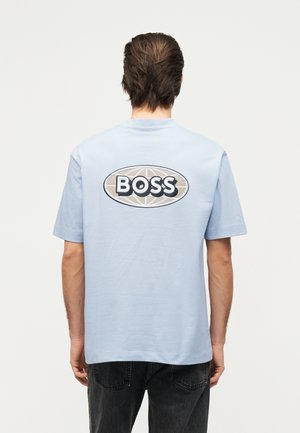 Osoba z krótkimi brązowymi włosami, ubrana w jasnoniebieską koszulkę z beżowo-białym logo "BOSS" na plecach, w połączeniu z czarnymi jeansami.