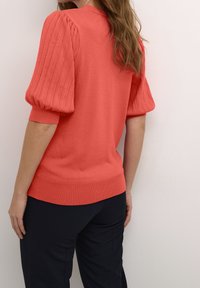 Frau mit welligem Haar, die einen korallfarbenen Sweater mit Puffärmeln und schwarze Hosen trägt, stehend vor einer hellen Wand, mit dem Rücken zur Kamera.