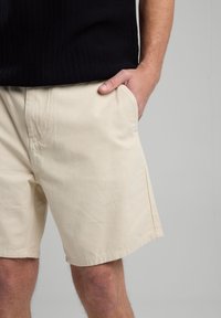 Pantaloni corti beige realizzati in tessuto leggero, con tasche laterali e vestibilità comoda. Il design include un orlo semplice senza motivi visibili.