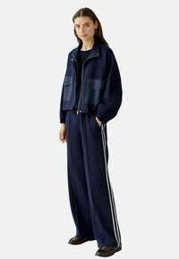 Veste zippée marine avec deux poches avant et pantalon large assorti avec des rayures blanches sur les côtés. Tissu lisse, coupe décontractée.
