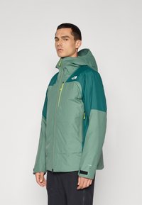 Veste pour hommes dans des tons de vert avec capuche, dotée d'une fermeture éclair à l'avant, de poignets ajustables et de contrastes en light vert. Matériau imperméable.