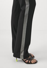 Black pants with a wide leg, featuring a vertical row of silver rhinestones along the side. Worn with black strappy sandals with a green accent.